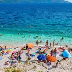 People enjoy a crowded pebble beach with colorful umbrellas by clear blue water, perfectly capturing the essence of summer in Croatia. Hills rise in the background, completing this picturesque scene that beckons travelers to experience Croatia's coastal charm.