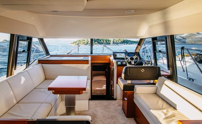Interior of a modern yacht showing white seating, a wooden table, steering wheel, and large windows with a view of water and distant hills.