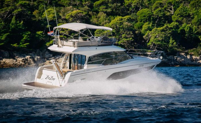 A white motor yacht speeds over the water near a forested shoreline, creating a wake behind it under a sunny sky.