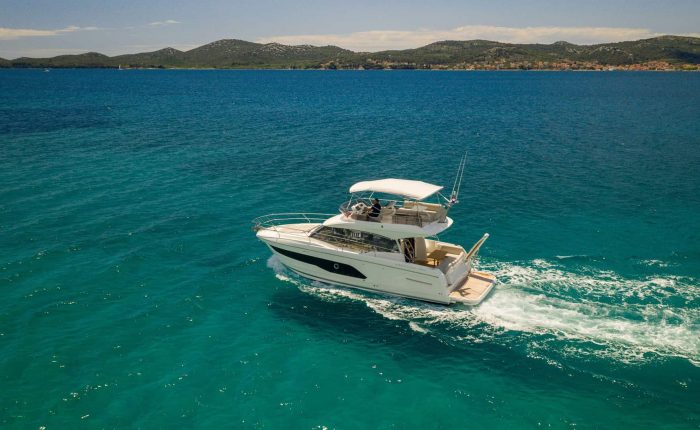 A white motor yacht travels across turquoise water with hills and a coastal town visible in the background under a partly cloudy sky.