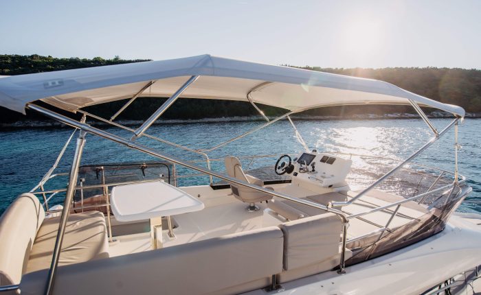 A sunlit view of a modern yacht's upper deck with shaded seating, a table, and a helm, set against a calm coastal backdrop.