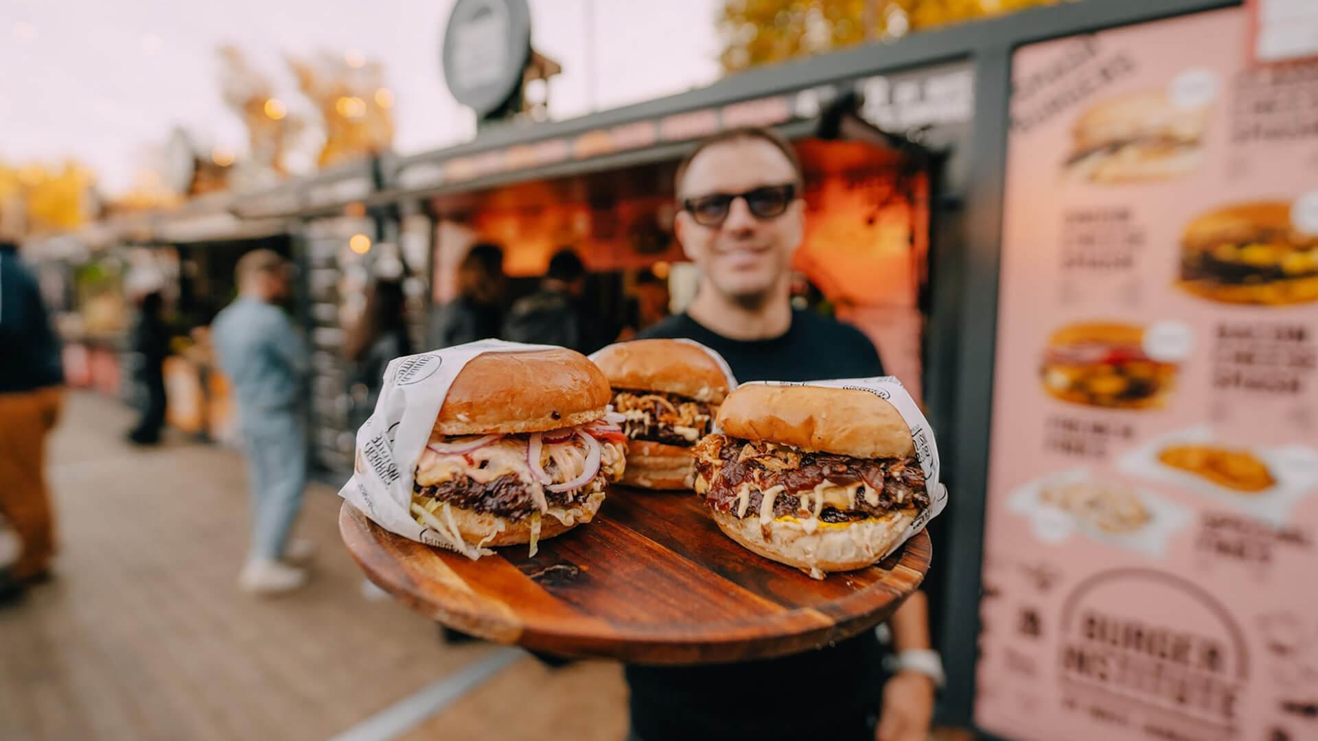 In June, a person holds a wooden platter with three loaded burgers in front of a Dubrovnik food stand displaying menu boards—one of the tastiest things to do in the city.