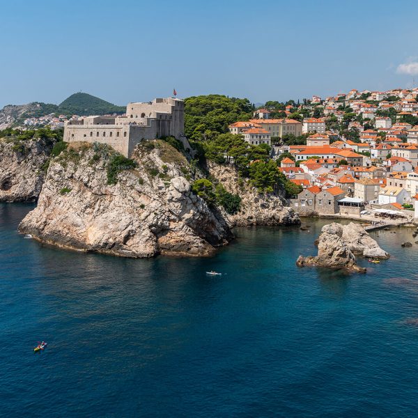 A coastal fortress atop rocky cliffs overlooks the Adriatic Sea, with a historic town of orange-roofed buildings clustered nearby.