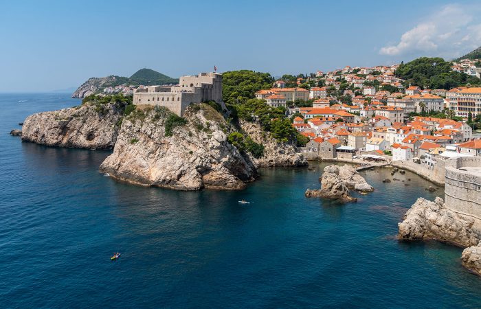A coastal fortress atop rocky cliffs overlooks the Adriatic Sea, with a historic town of orange-roofed buildings clustered nearby.