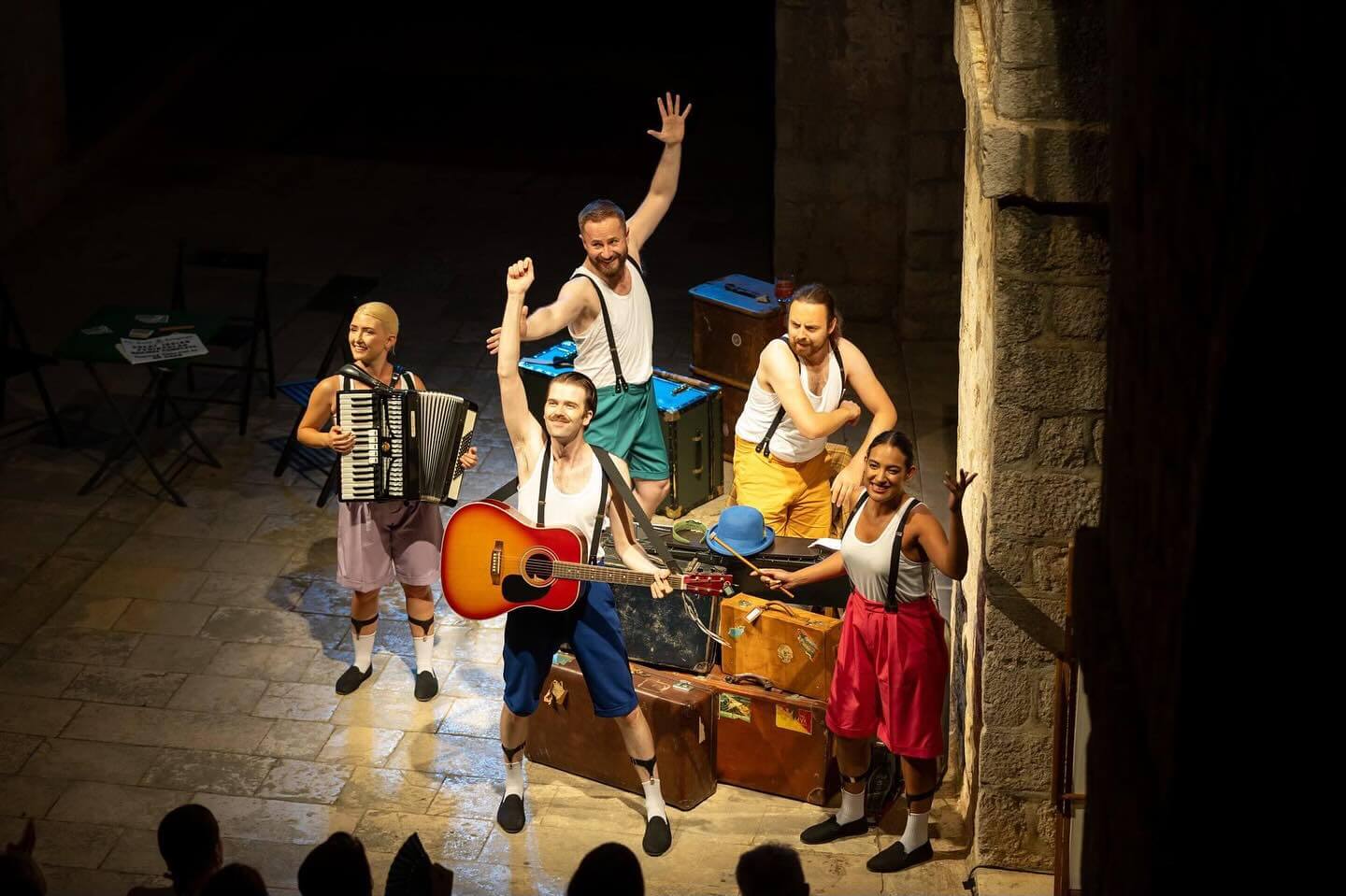 Five performers in colorful costumes stand on a stone stage in Dubrovnik, holding musical instruments and smiling at an audience, with suitcases and props behind them—an unmissable highlight among June events and things to do in the city.