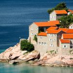 Stone buildings with orange tile roofs sit on a rocky coastline next to clear blue water, with trees and greenery in the background—a view often admired on Croatia tours.