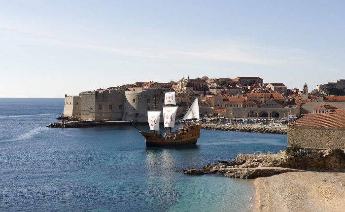A large wooden sailing ship with white sails approaches the medieval stone walls of a coastal city under a clear sky, with calm blue water and sandy beach in the foreground.