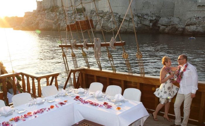 A couple stands on the deck of a docked boat decorated with flowers, beside a set table, with a medieval stone wall and water in the background at sunset.
