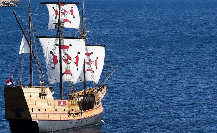 A wooden sailing ship with three masts and white sails marked by red and black symbols floats on calm blue sea.