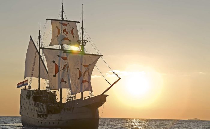 A large sailing ship with decorated sails travels on the ocean at sunset, with the sun low on the horizon and a small island visible in the distance.