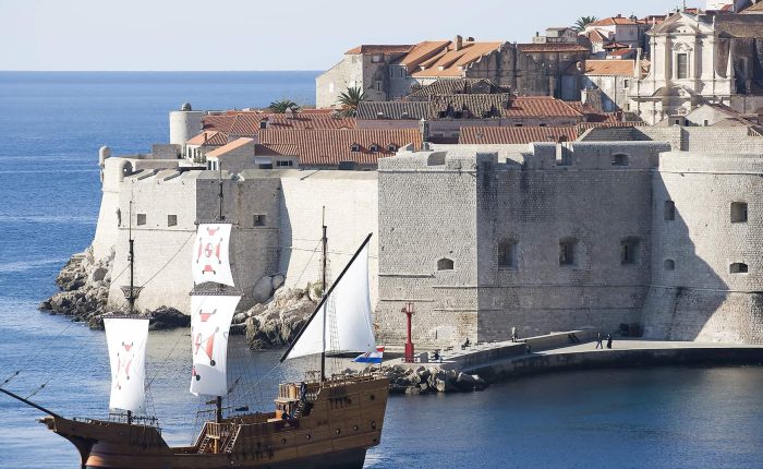 A wooden sailing ship with white sails passes near the stone walls and red-roofed buildings of a coastal fortress city by the sea.
