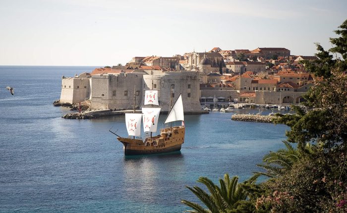 A wooden sailing ship with white sails is near the historic stone walls and red-roofed buildings of Dubrovnik, Croatia, with calm blue water and greenery in the foreground.