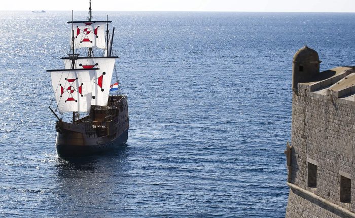 A wooden sailing ship with cross-adorned sails is on the sea near a stone fortification under a clear sky.