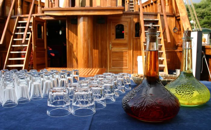 A table covered with a blue cloth holds rows of empty glasses and two glass decanters, one filled with red liquid and the other with yellow, on the deck of a wooden boat.