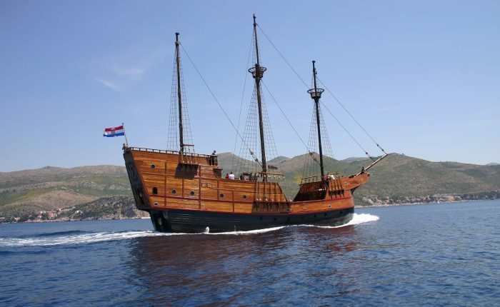 A wooden sailing ship with two masts and a Croatian flag moves across calm blue water with hills in the background.