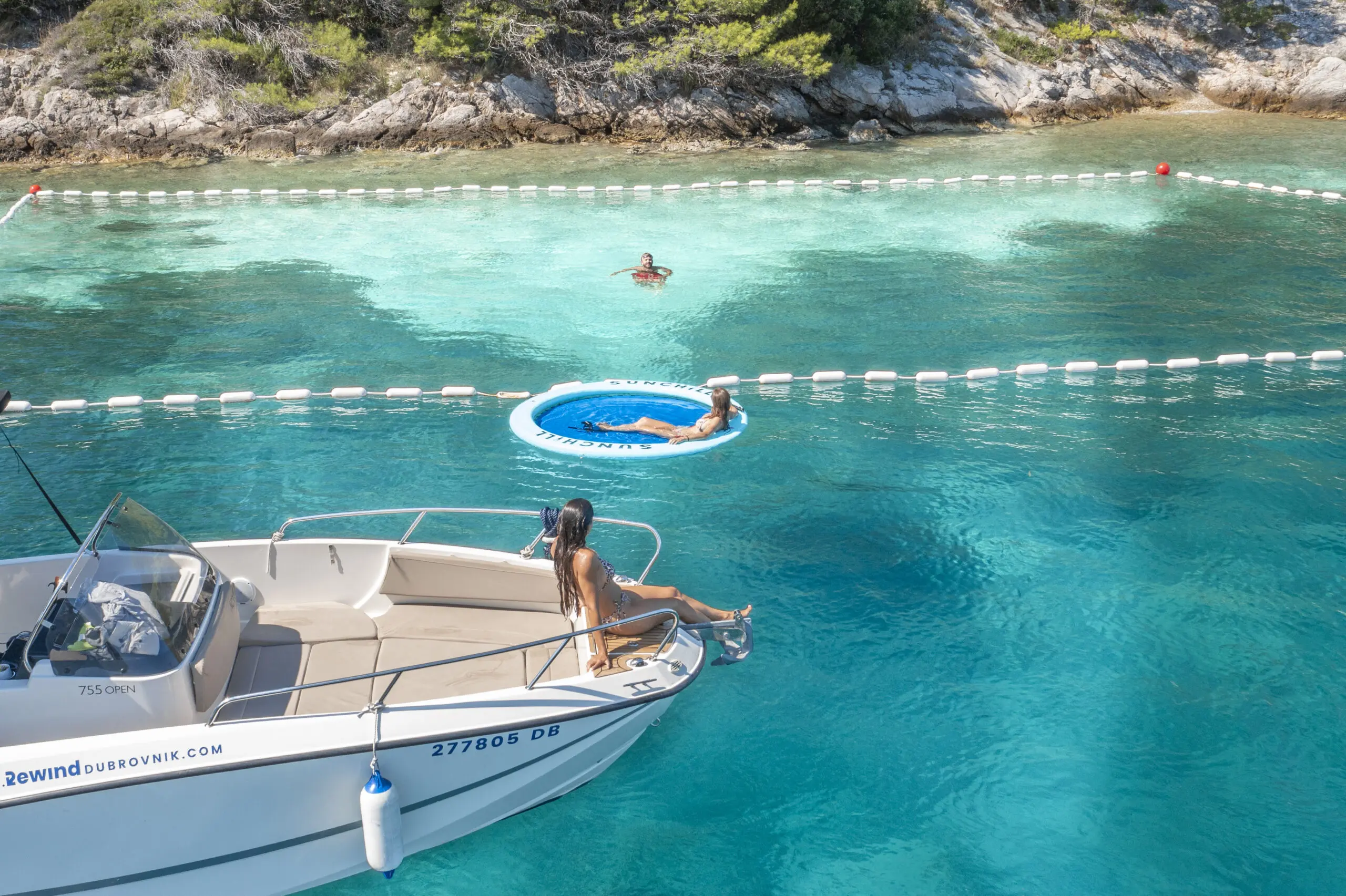 A woman sits on a boat while another person floats on a blue inflatable ring in a roped-off swimming area of clear turquoise water, soaking in the scenic beauty often explored on Dubrovnik Tours near the rocky, tree-lined shore.