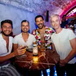 Four men sit around a small round table with drinks in a dimly lit bar, colorful neon lighting casting a glow—reminiscing about their ultimate romantic day on the Adriatic Sea, surrounded by shelves of bottles in the background.