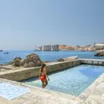 A woman in a red swimsuit walks into a seaside pool with stone surroundings, overlooking the sea and an old town with orange rooftops—a scene straight from the Best LGBTQ Travel Guide to Croatia.