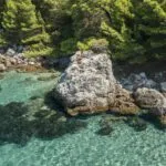 A person sits on a large rock beside clear turquoise water as another floats on an inflatable near a rocky, tree-lined shore—an idyllic scene perfect for a Dubrovnik private day boat adventure. A person sits on a large rock beside clear turquoise water as another floats on an inflatable near a rocky, tree-lined shore—an idyllic scene perfect for a Dubrovnik private day boat adventure.
