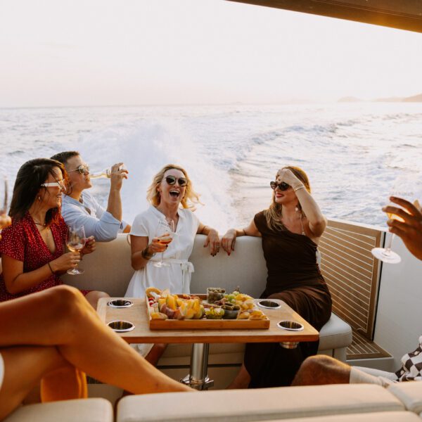 Six people sit on a boat, smiling and holding drinks around a table with snacks, with the sea and coastline in the background—discover Dubrovnik and its boat charters for unforgettable moments like these.