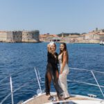 Two women stand on the bow of a boat, posing for a photo with Dubrovnik’s historic stone fortifications in the background—a perfect moment from the Travel Agents’ Guide: Private Boat Tours in Dubrovnik.
